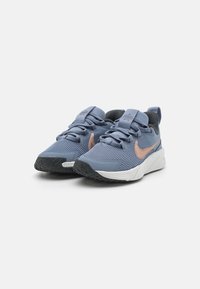 Kék sportcipők texturált szövet felsőrésszel, rózsaszín swoosh logóval, fűzős záródással és fekete gumi talppal. A fehér középtalpa párnázottságot biztosít.