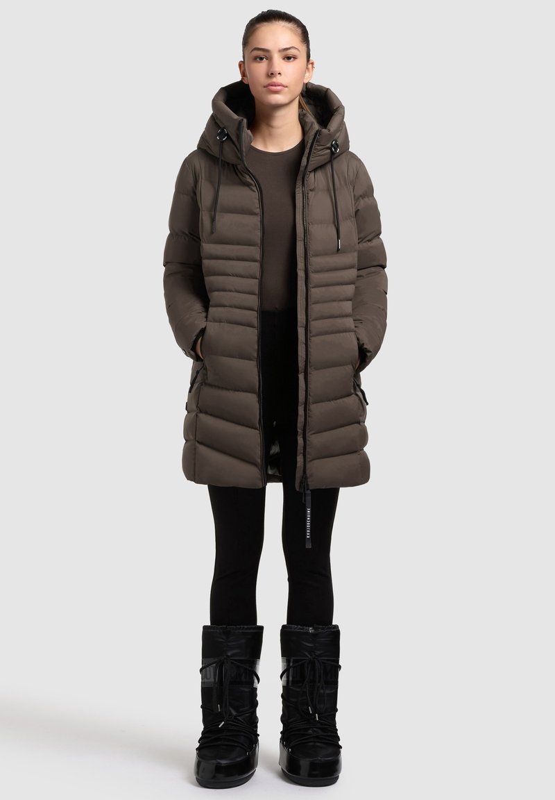 Abrigo acolchado marrón con capucha, con cierre de cremallera en la parte delantera, bolsillos laterales y un diseño acolchado. Combinado con botas negras, impermeables y hasta la rodilla.