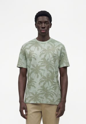 Jonge man met een lichtgroen T-shirt met palmboompatronen en beige broek, staand tegen een effen witte achtergrond.