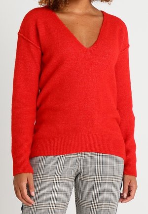 Kobieta ubrana w czerwony sweter z dekoltem w serek i długimi rękawami oraz czarno-białe spodnie w kratę, stoi z ramionami swobodnie opuszczonymi wzdłuż ciała.