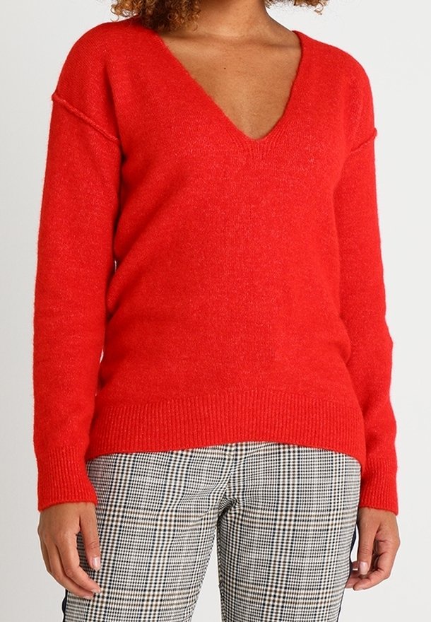 Femme portant un pull rouge à manches longues avec un col en V et un pantalon à carreaux noir et blanc, debout, les bras détendus le long du corps.