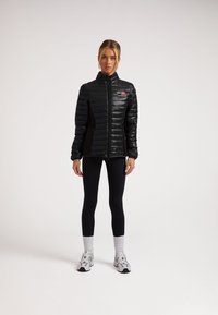 Giacca a vento nera con colletto alto e zip frontale, pannelli verticali cuciti e un piccolo logo rosso sul lato sinistro del petto. Indossata sopra leggings neri.