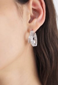 Silver hoop earrings featuring a textured design, with layered accents and an oval shape, worn on a person's ear.
