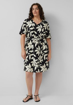 Femme aux cheveux bouclés portant une robe noire mi-longue avec un grand motif floral blanc et des sandales noires, debout devant un fond gris.