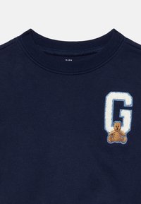 Marineblå babygenser med ribbet rundhalset og brodert hvitt bokstav "G" ved siden av et lite brunt kosedyr-patch.