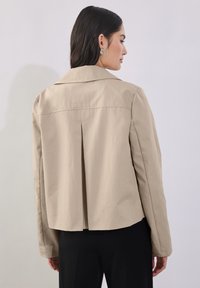 Beige lyhyt takki, jossa on jämäkkä kaulus ja takana laskostus. Valmistettu sileästä kankaasta, jossa on pitkät hihat ja minimiaaliset yksityiskohdat.