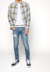 Camisa de franela a cuadros en azul y verde oliva, camiseta blanca, vaqueros azules desgastados y zapatillas verdes con detalles y cordones blancos.