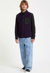 Lila Fleeceweste mit hohem Kragen und durchgehendem Reißverschluss, ausgestattet mit einer schwarzen Brusttasche. Trägt man über einem schwarzen Langarmshirt und hellen Jeans.