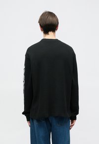 Μαύρο πουκάμισο με πλέξη waffle, μακρύ μανίκι, στρογγυλή λαιμόκοψη, και ρίγες γραφικών σε αντίθεση κατά μήκος του αριστερού μανικιού.