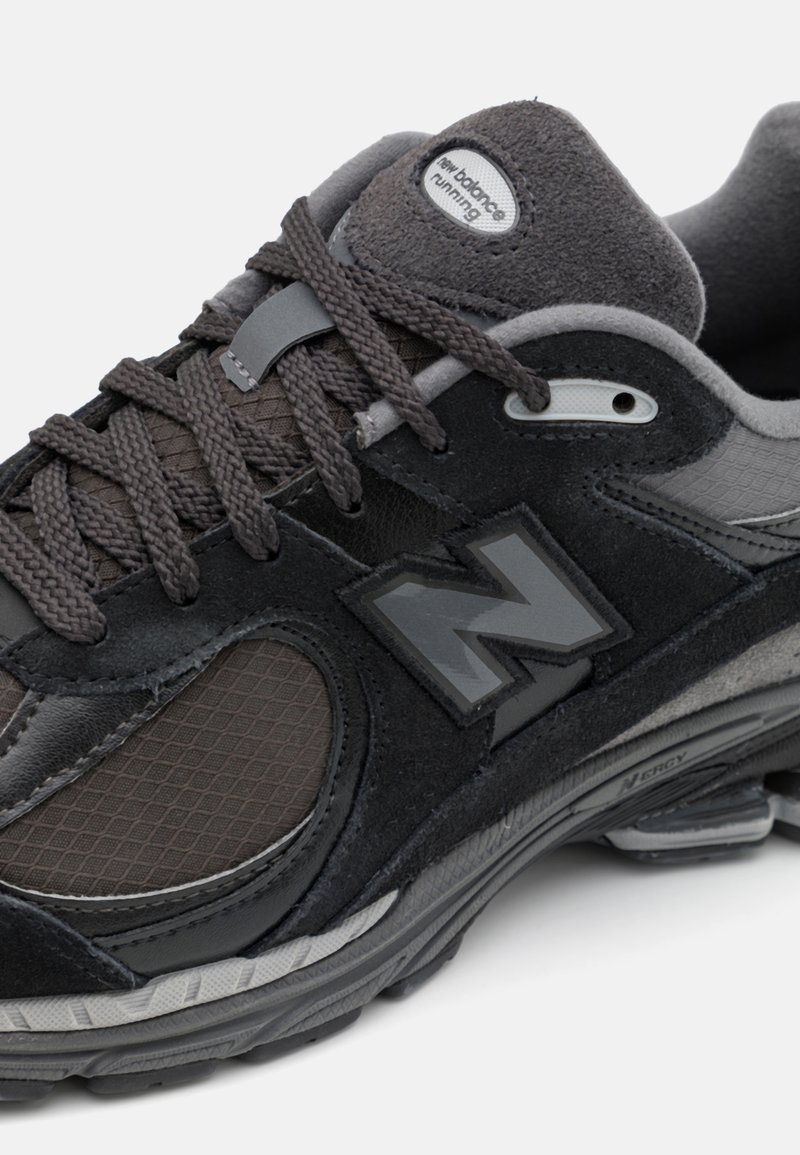Zwarte en grijze sportschoen met mesh- en suède-accenten, voorzien van gestructureerde veters, een gevoerde rand en een opvallend "N"-logo op de zijkant.