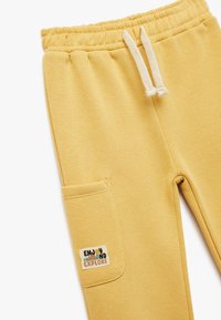 Pantalones de chándal amarillos hechos de una tela suave, con una cinturilla elástica, cordones blancos y un bolsillo con parche que lleva una etiqueta que dice "Disfruta y Explora".