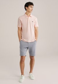 Svetloružová polo tričko s klasickým golierom, skombinované so svetlomodrými kockovanými šortkami. Model má na sebe biele tenisky a stojí s rukami v vreckách.