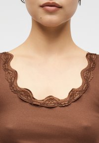 Κοντινή λήψη στο λαιμό και το πάνω μέρος του στήθους ενός ατόμου που φοράει καφέ ριμπ πλεκτό τοπ με διακοσμητική δαντέλα με λουλούδια κατά μήκος του λαιμού.