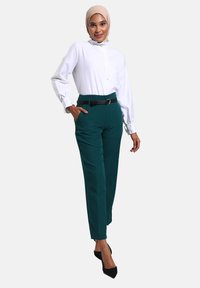 Blouse blanche avec col montant et détails de boutons, associée à un pantalon vert sur mesure et une ceinture noire, portée avec des escarpins noirs à bout pointu.