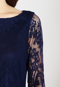Marinblå klänning med en solid överdel och genomskinliga långärmade spetsärmar med blommönster och rundad halsringning.