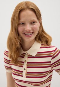 Mujer joven con cabello rojo sonriendo, vestida con una camisa de polo a rayas con líneas horizontales rojas, beige y marrones sobre un fondo liso.