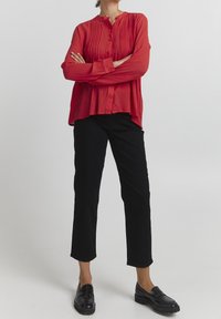 Blusa roja de manga larga con pliegues verticales, escote redondo y cierre con botones, combinada con pantalones negros cortos y mocasines negros.