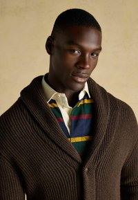 Barna kötött kardigán sálgallérral, multiszínű csíkos pólóing felett viselve. A textúra bordázott, jól látható varrási részletekkel.