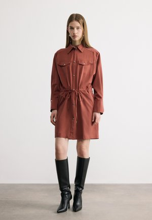 Ženska oblečena v rjavo rjavo srajčno obleko z dolgimi rokavi in sprednjimi žepi ter črne škornje s petami do kolen, stoji ob enobarvnem ozadju.