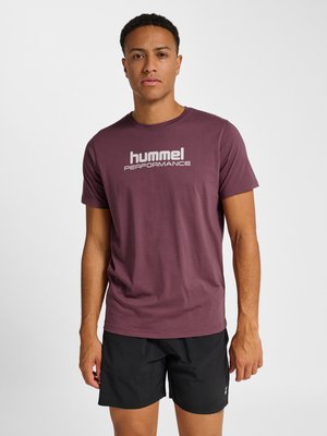 Mężczyzna ubrany w fioletowy t-shirt "hummel performance" i czarne szorty, stojący na jednolitym białym tle.