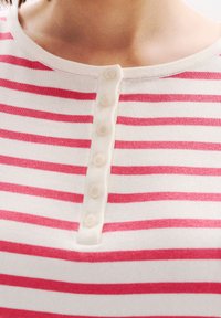 Raidallinen paita, jossa on vuorottelevia valkoisia ja vaaleanpunaisia raitoja, pyöreä kaula-aukko ja kuusi pientä nappia etupaneelissa. Pehmeä kangas.