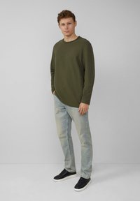 Olijfgroene langwerpige shirt met ronde hals, gecombineerd met lichtblauwe jeans en zwarte sneakers. Vlakke textuur, relaxte pasvorm en geen zichtbare patronen.