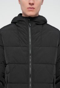 Black padded jacket with a hood, featuring a front zipper and quilted texture. The material appears lightweight and smooth.