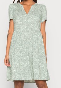 Femme portant une robe vert clair à manches courtes, longueur genou, avec un motif de feuilles blanches et un décolleté en V, montrant des tatouages sur les bras.