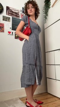 Osoba na sebe má vzorované midi šaty, červené slingback topánky a drží malú červenú kabelku na rameno, stojí vo vnútri pri bielej komode.