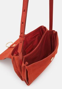 Oranje suède schoudertas met een enkele verstelbare schouderband, meerdere binnencompartimenten en een sluiting met gouden knop. Gestructureerd oppervlak.