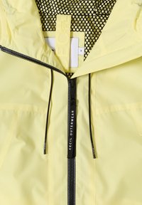 Veste jaune clair avec fermeture éclair noire et cordons, doublure intérieure en mesh noir et jaune, et une étiquette de taille marquée "S" à l'intérieur du col.