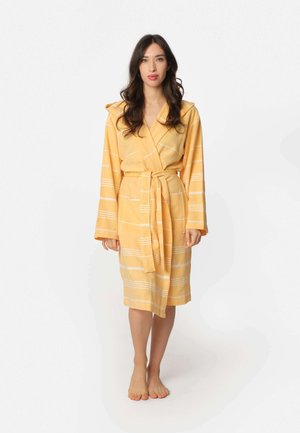 Woman standing barefoot wearing a yellow hooded bathrobe with white horizontal stripes tied at the waist against a plain background.