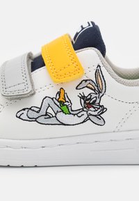 Sneaker bianca con tomaia testurizzata, cinturini in velcro grigi e gialli. Presenta un personaggio dei cartoni ricamato con una carota nei colori verde e arancione.