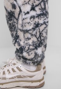 Fleece joggers in black and beige pattern with textured finish. Features a side zipper and embroidered detail. Paired with beige sneakers.