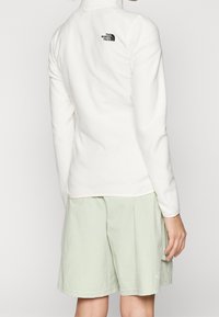 Pullover crema con colletto alto e dettaglio logo nero sulla schiena, abbinato a pantaloni corti verde chiaro con tasca laterale.