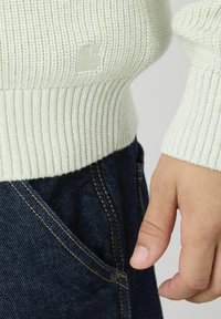 Pull en maille côtelée vert clair avec une taille cintrée, présentant un petit détail de logo. Associé à un jean en denim noir, montrant la poche.