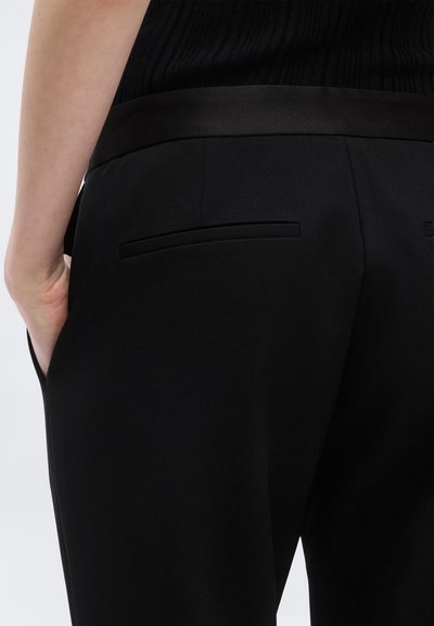 Personne portant un pantalon noir sur mesure avec un haut côtelé noir, la main reposant dans la poche latérale, montrant les détails de la taille arrière et de la poche.