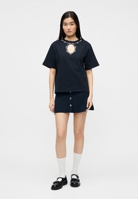 Νέα γυναίκα που φοράει μπλε ναυτικό μπλουζάκι με κόσμημα στο ντεκολτέ σε σχήμα κλειδαρότρυπας, μπλε ναυτική φούστα με κουμπιά, λευκές κάλτσες και μαύρα παπούτσια Mary Jane, στέκεται μπροστά σε λευκό φόντο.