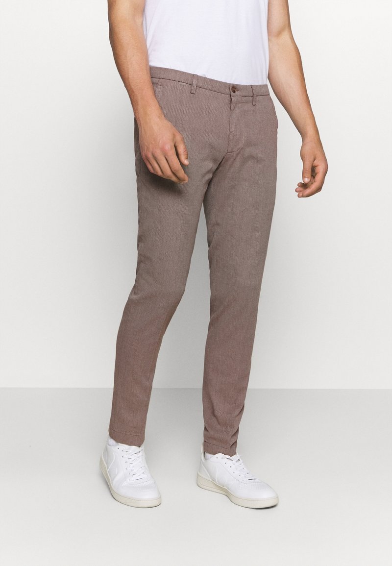 Homme portant un pantalon slim marron clair, un t-shirt blanc et des baskets blanches, debout devant un fond clair et uni.