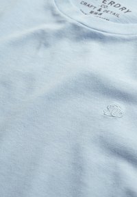 Lichtblauw katoenen T-shirt met een ronde hals en een klein geborduurd logo aan de voorkant links. Vloeiende textuur en minimalistisch ontwerp.