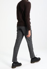 Brązowy sweter z dzianiny, zwężane szare spodnie w kratę i czarne sznurowane buty z grubą podeszwą, widok z tyłu.