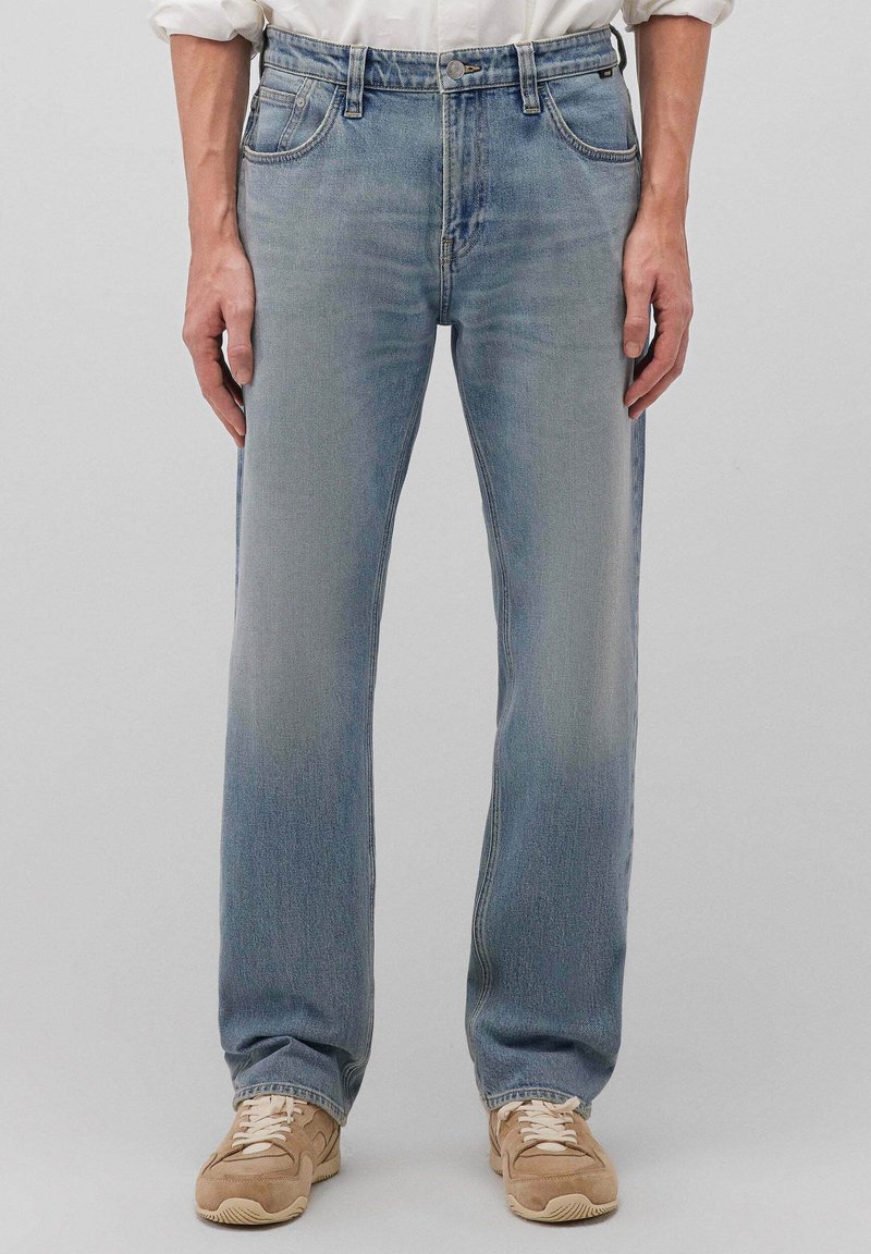 Persona che indossa jeans azzurri a gamba dritta, camicia bianca con le maniche rimboccate e sneakers beige, in piedi davanti a uno sfondo chiaro uniforme.
