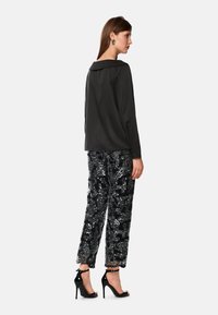 Frau mit schwarzem langärmligem Oberteil, schwarzen und silbernen Pailletten-Hosen, schwarzen hohen Sandalen und großen schwarzen Ohrringen, die seitlich steht.