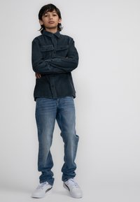 Tmavě modrá kordrová košile s náprsními kapsami, v kombinaci se světle modrými džínami a bílými teniskami s černými detaily.