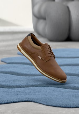 Chaussure marron à lacets en cuir avec doublure en chaussette beige flottant au-dessus d'un tapis bleu texturé, mobilier rembourré gris en arrière-plan.