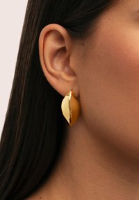 Primo piano dell'orecchio di una donna con lunghi capelli scuri che indossa un orecchino a lobo di forma fogliare, liscio e lucente, in oro.