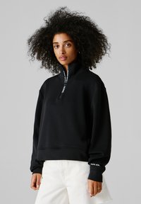 Ženska s kodrastimi lasmi, oblečena v črni pulover z visokim ovratnikom z zadrgo in belimi hlačami, stoji na enostavnem sivem ozadju.