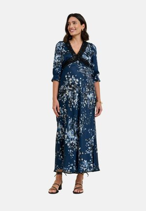 Lachende vrouw die een lange marineblauwe bloemenjurk draagt met zwarte kanten rand en zwarte sandalen met bandjes, staand tegen een effen achtergrond.