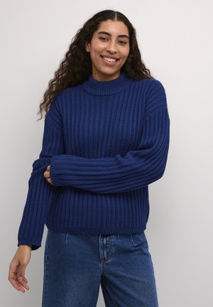 Pulover ribbed de culoare bleumarin, cu gât înalt și mâneci lungi, asortat cu blugi din denim cu talie înaltă. Țesătura are o textură tricotată.