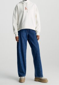 Felpa crema oversize con logo, abbinata a jeans in denim blu scuro a gamba larga. Calzature chunky beige-marroni, texture morbide e design casual.